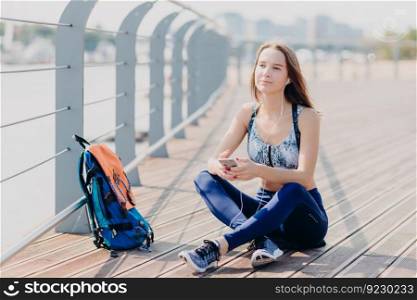 People and active lifestyle concept. Attractive young female enjoys calm atmosphere in urban territory, breathes fresh air, sits crossed legs, listens favourite music from playlist, uses mobile phone