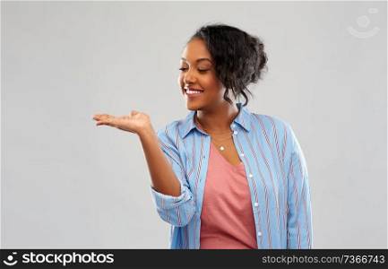 people and advertisement concept - happy african american young woman holding something imaginary on empty hand over pink background. happy african woman holding something on hand