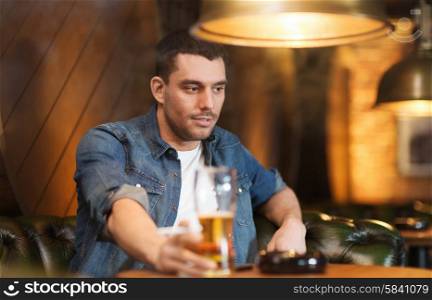 people and bad habits concept - young man drinking beer and smoking cigarette at bar or pub. man drinking beer and smoking cigarette at bar