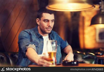 people and bad habits concept - young man drinking beer and smoking cigarette at bar or pub. man drinking beer and smoking cigarette at bar