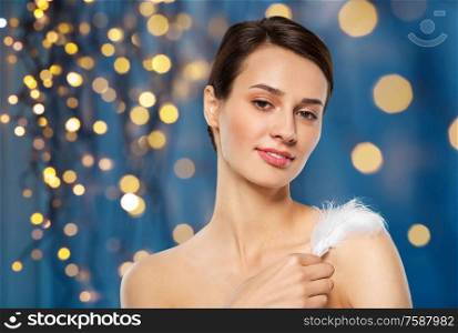 people and beauty concept - beautiful woman with feather touching her shoulder skin over holidays lights on dark blue background. beautiful woman with feather touching her shoulder