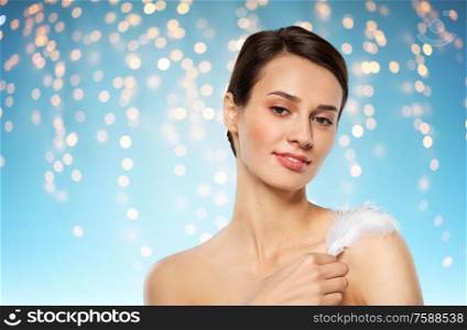 people and beauty concept - beautiful woman with feather touching her shoulder skin over holidays lights on blue background. beautiful woman with feather touching her shoulder