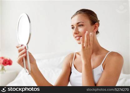 people and beauty concept - beautiful woman with mirror sitting on bed at home bedroom and touching her face skin. beautiful woman with mirror sitting on bed at home