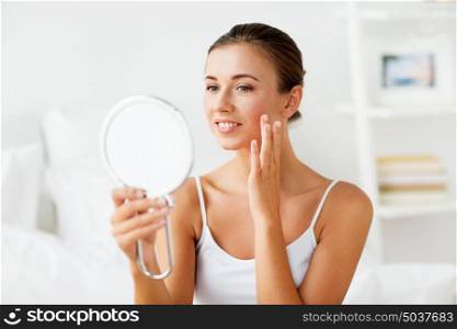 people and beauty concept - beautiful woman with mirror touching her face skin at home bedroom. beautiful woman with mirror touching her face skin