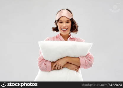 people and bedtime concept - happy smiling young woman in pajama and eye sleeping mask hugging pillow over grey background. woman with pillow in pajama and eye sleeping mask