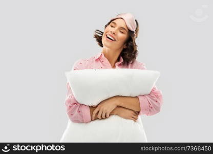 people and bedtime concept - happy young woman in pajama and eye sleeping mask hugging pillow over grey background. woman with pillow in pajama and eye sleeping mask