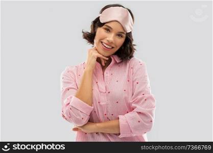 people and bedtime concept - happy young woman in pajama and eye sleeping mask over grey background. happy young woman in pajama and eye sleeping mask