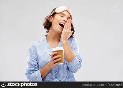people and bedtime concept - happy young woman in pajama and eye sleeping mask with takeaway paper cup of coffee yawning over grey background. woman in pajama and eye mask with coffee yawning
