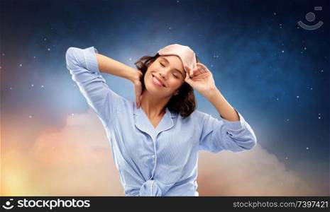 people and bedtime concept - happy young woman in pajama and eye sleeping mask over starry night sky and cloud background. happy young woman in pajama and eye sleeping mask