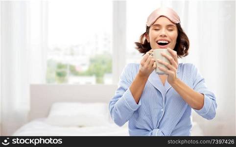 people and bedtime concept - happy young woman in pajama and eye sleeping mask drinking coffee from mug over background. woman in pajama and sleeping mask drinking coffee