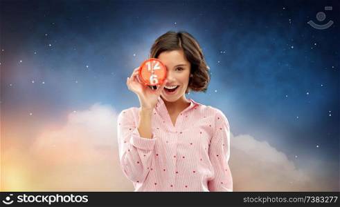 people and bedtime concept - happy young woman in pajama covering one eye with alarm clock showing six o&rsquo;clock over night sky background. happy young woman in pajama with alarm clock