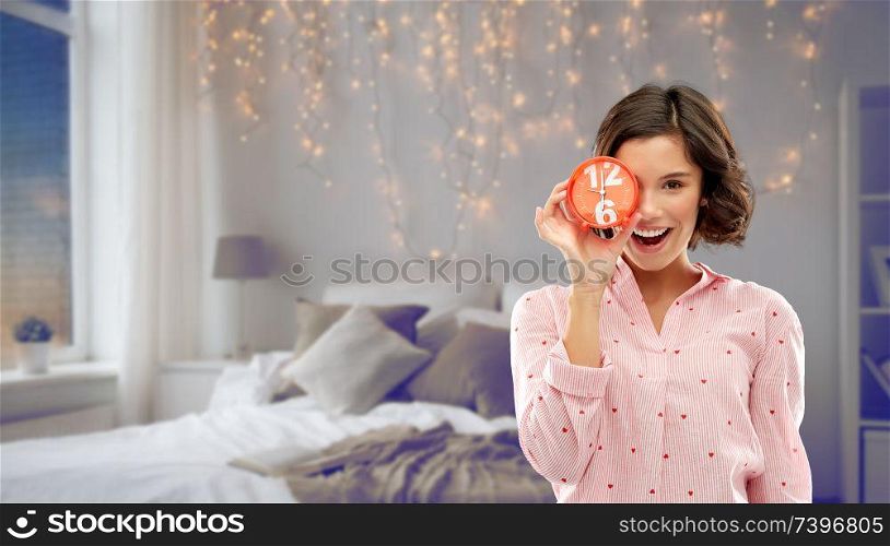 people and bedtime concept - happy young woman in pajama covering one eye with alarm clock over bed and garland lights at night bedroom background. happy young woman in pajama with alarm clock