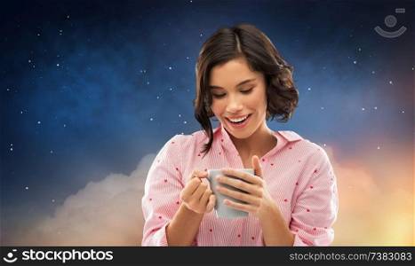 people and bedtime concept - happy young woman in pajama with mug of coffee over starry night sky background. happy young woman in pajama with mug of coffee
