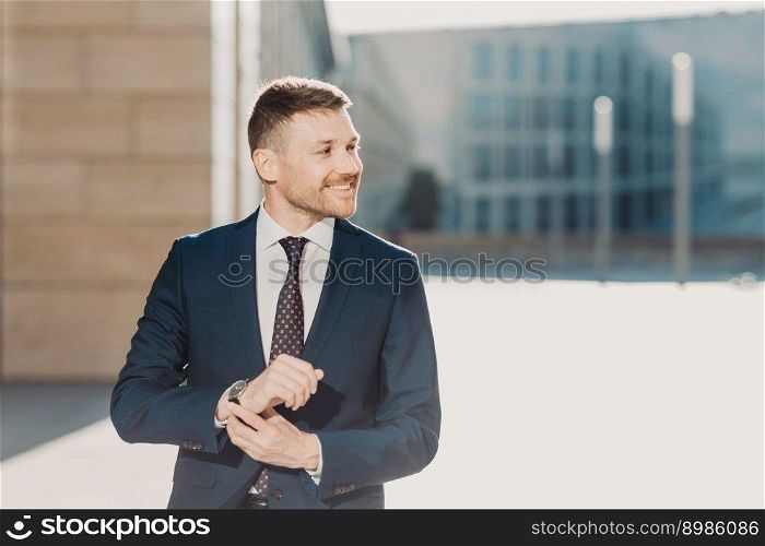 People and business concept. Glad elegant young businessman with stubble, wears formal clothes and watch, waits for someone, poses outdoor near office. People, career and leadership concept.