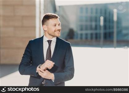 People and business concept. Glad elegant young businessman with stubble, wears formal clothes and watch, waits for someone, poses outdoor near office. People, career and leadership concept.