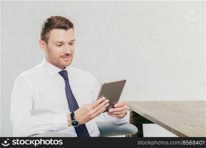 People and business concept. Young businessman in formal outfit uses touch pad for reading financial news in internet, connected to wireless internet in office, poses against white wall. Man banker
