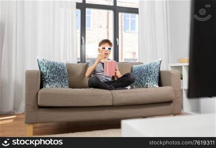 people and childhood concept - boy in 3d movie glasses eating popcorn from striped bucket and watching tv at home. boy in 3d movie glasses and watching tv at home