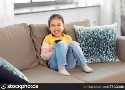 people and childhood concept - happy smiling little girl with smartphone sitting on sofa at home. happy smiling little girl with smartphone at home