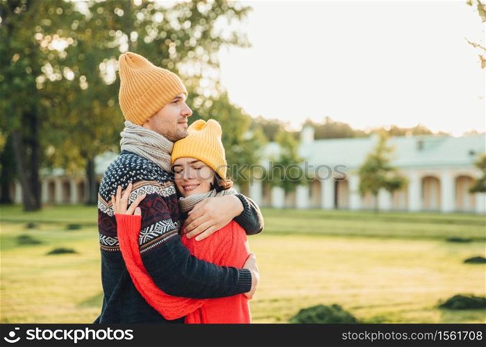 People and closeness concept. Young couple in love have date, embrace each other, feel support, being alone in park, have perfect relationships. Handsome man in warm sweater and hat hugs girlfriend