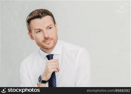 People and confidence concept. Attractive young unshaven male employee looks thoughtfully aside, contemplates about something important, wears white shirt and tie, isolated over white background