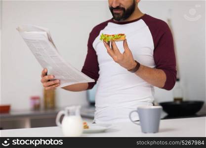 people and diet concept - man eating sandwich with coffee for breakfast and reading newspaper at home kitchen. man reading newspaper and eating at home kitchen