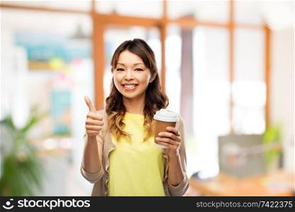 people and drinks concept - happy young asian woman drinking takeaway coffee from paper cup and showing thumbs up over office background. asian woman drinking coffee and showing thumbs up