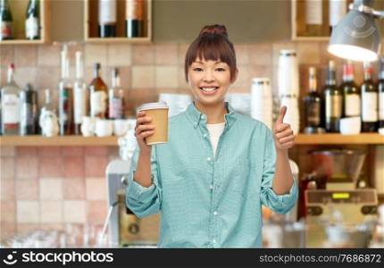 people and drinks concept - happy young asian woman with takeaway coffee in paper cup showing thumbs up over bar background. asian woman with coffee showing thumbs up at bar