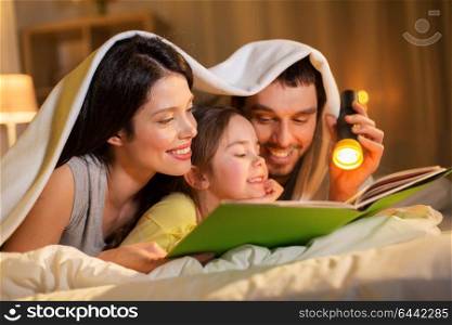 people and family concept - happy mother, father and little daughter reading book with torch light in bed lying under blanket at night at home. happy family reading book in bed at night at home
