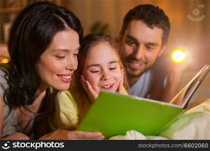 people and family concept - happy mother, father and little daughter reading book with torch light in bed at night at home. happy family reading book in bed at night at home