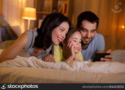 people and family concept - happy mother, father and little daughter with smartphone in bed at night at home. happy family with smartphone in bed at night