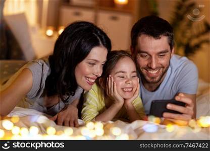 people and family concept - happy mother, father and little daughter with smartphone in bed at night at home. happy family with smartphone in bed at night