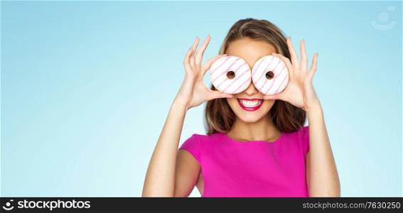 people and fast food concept - happy young woman or teen girl in pink dress having fun and looking through donuts over blue background. happy woman or teen girl looking through donuts