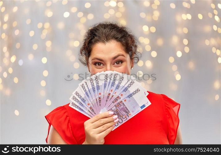 people and finances concept - happy woman in red dress hiding her face behind euro money banknotes over festive lights background. happy woman holding euro money banknotes