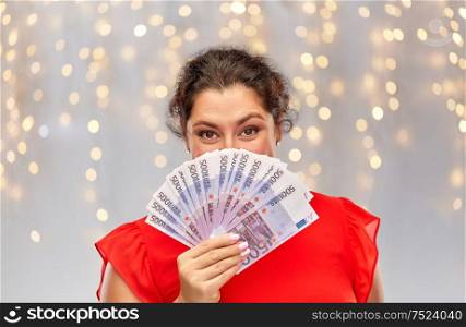 people and finances concept - happy woman in red dress hiding her face behind euro money banknotes over festive lights background. happy woman holding euro money banknotes