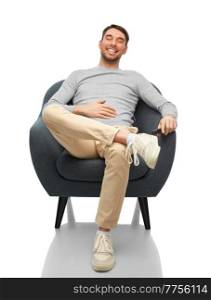 people and furniture concept - happy smiling man sitting in chair and touching his tummy over white background. happy smiling young man sitting in chair