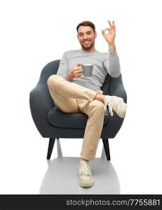 people and furniture concept - happy smiling man with cup of coffee or tea sitting in chair and showing ok gesture over white background. man with mug showing ok gesture sitting in chair