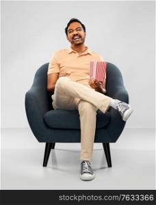 people and furniture concept - happy smiling young indian man eating popcorn sitting in chair over grey background. happy smiling indian man eating popcorn in chair