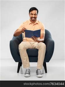 people and furniture concept - happy smiling young indian man sitting in chair and reading book over grey background. happy young indian man reading book in chair