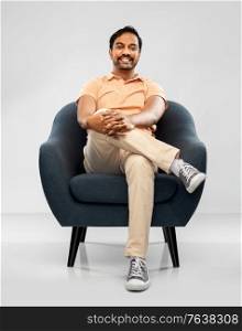 people and furniture concept - happy smiling young indian man sitting in chair over grey background. happy smiling young indian man sitting in chair
