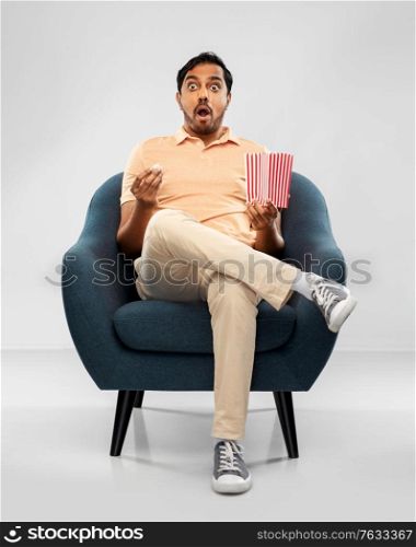 people and furniture concept - shocked young indian man eating popcorn sitting in chair over grey background. shocked indian man eating popcorn in chair