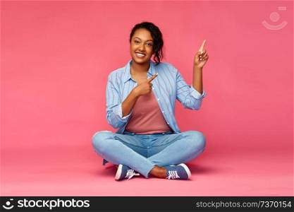 people and gesture concept - happy african american young woman pointing fingers up and sitting on floor over pink background. happy african american woman pointing fingers up