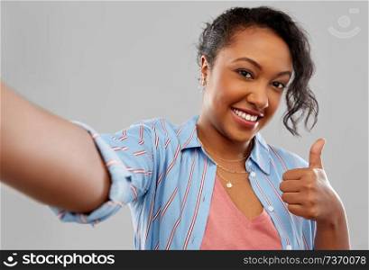 people and gesture concept - happy african american young woman taking selfie and showing thumbs up over grey background. happy african american woman taking selfie