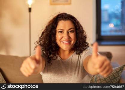 people and gesture concept - happy smiling young woman showing thumbs up on sofa at home. happy smiling woman showing thumbs up at home