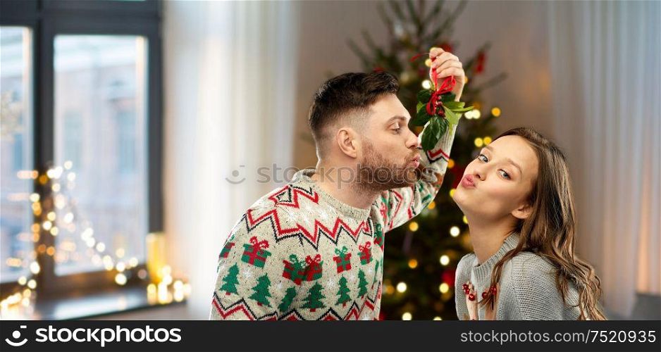 people and holiday traditions concept - portrait of happy couple in ugly sweaters kissing under mistletoe over home and christmas tree lights on background. happy couple kissing under mistletoe on christmas