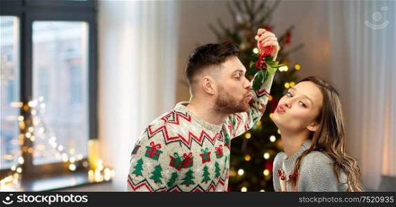 people and holiday traditions concept - portrait of happy couple in ugly sweaters kissing under mistletoe over home and christmas tree lights on background. happy couple kissing under mistletoe on christmas