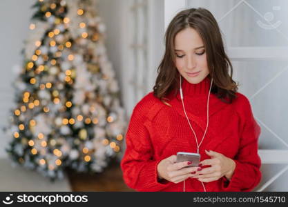 People and holidays concept. Concentrated dark haired young woman holds cell phone in hands, reads news in internet, uses earphones, stands indoor near fir tree, connected to high speed internet