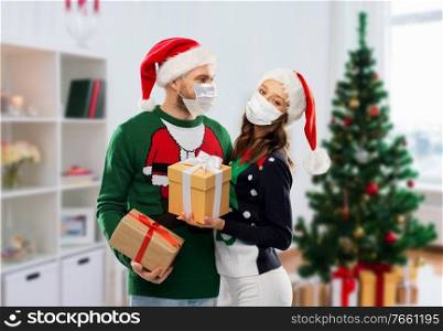 people and holidays concept - couple in santa hats and sweaters wearing face protective medical mask for protection from virus disease with christmas gifts at home. couple in masks, christmas sweaters with gifts