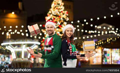 people and holidays concept - happy couple in santa hats and ugly sweaters with gifts over christmas market lights background. happy couple with gifts at christmas market