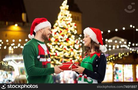 people and holidays concept - happy couple in santa hats and ugly sweaters with gift box over christmas market lights background. happy couple with gift at christmas market