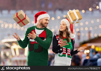 people and holidays concept - happy couple in santa hats and ugly sweaters with gifts over christmas market background. happy couple in christmas sweaters with gifts